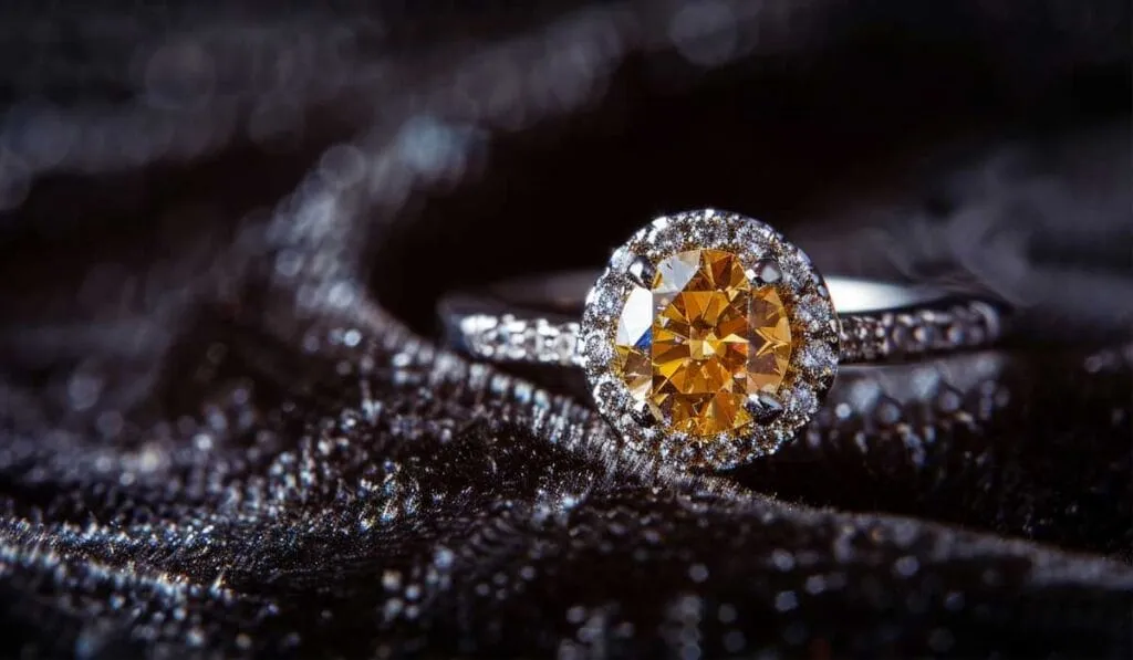 A close-up of a diamond ring featuring an orange center stone surrounded by smaller white diamonds, placed on a dark, textured fabric background.