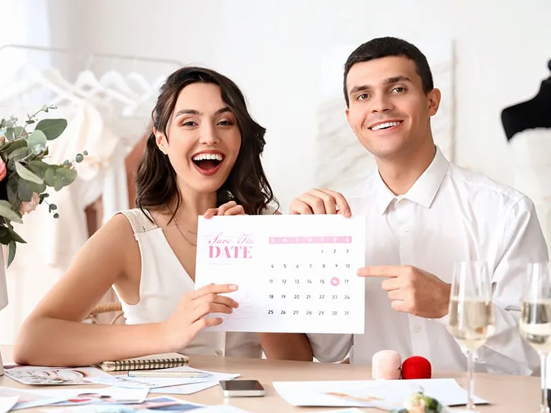 A smiling woman and man hold up a "Save the Date" calendar for their wedding, pointing to a specific date. Various wedding planning items are on the table in front of them.