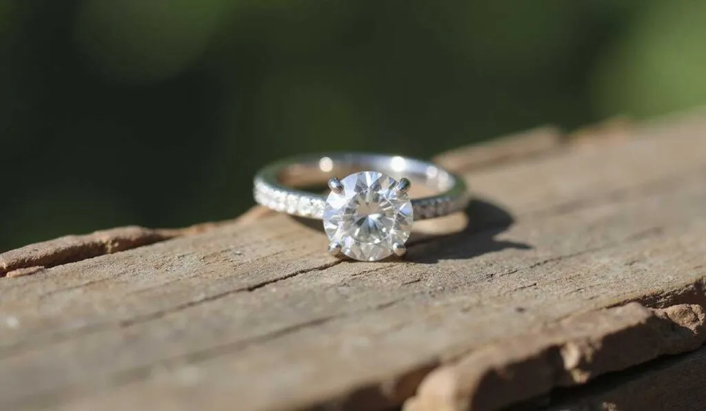 A diamond engagement ring with a round-cut center stone and a band featuring smaller diamonds rests on a weathered wooden surface.